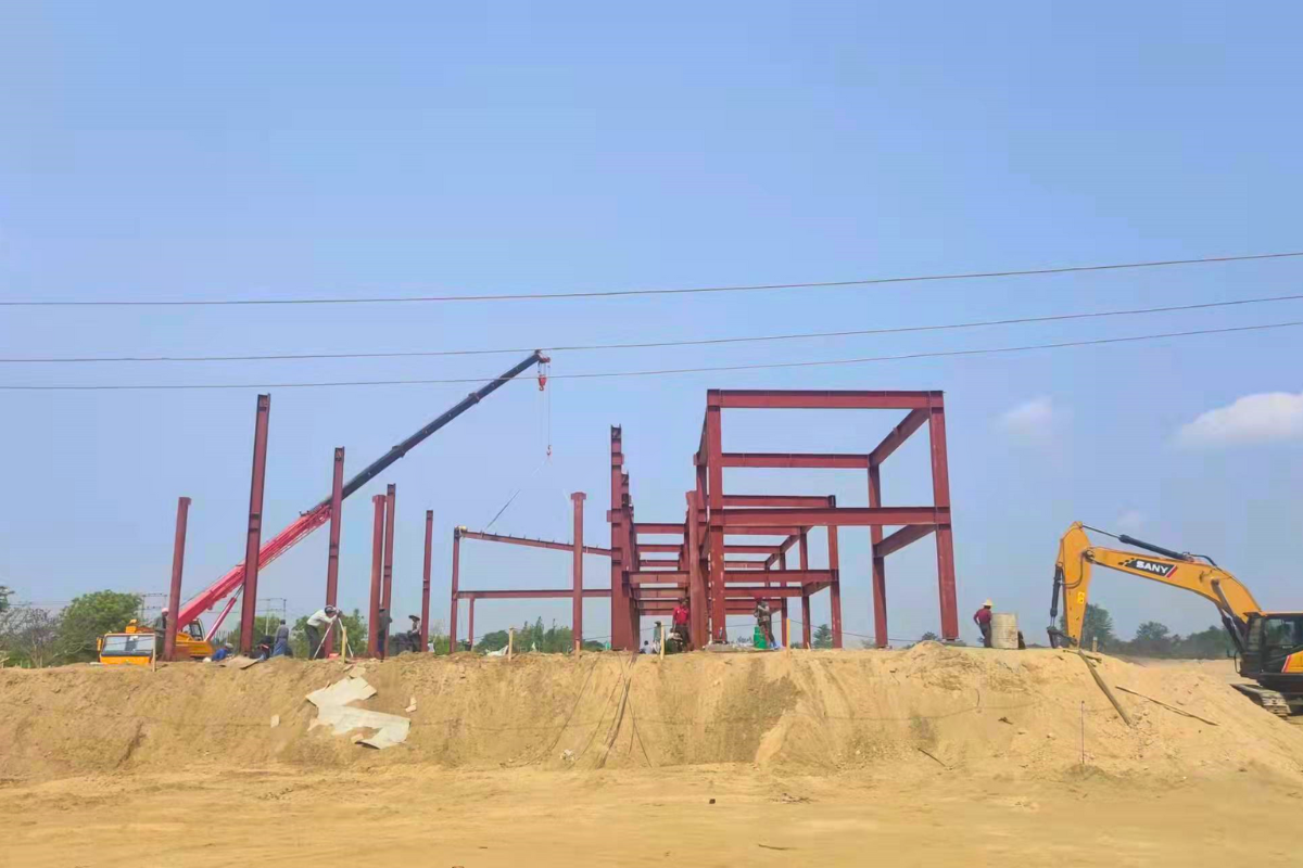 Early stages of steel structure erection for Emerald E-Commerce Center in Naypyidaw, Myanmar, showing red steel columns being placed with crane assistance.