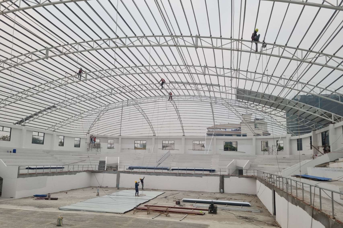 Construction workers on the arched steel roof structure during installation at Ba Htoo Indoor Sport Stadium in Mandalay, Myanmar, showing progress of the super structure.