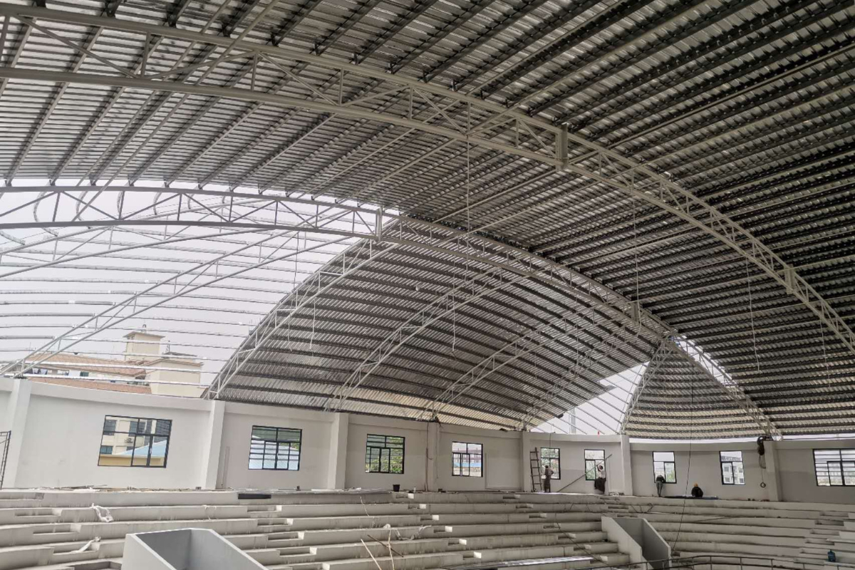 Installation of roof sheeting onto the curved steel structure at Ba Htoo Indoor Sport Stadium in Mandalay, Myanmar, nearing completion of the roof.