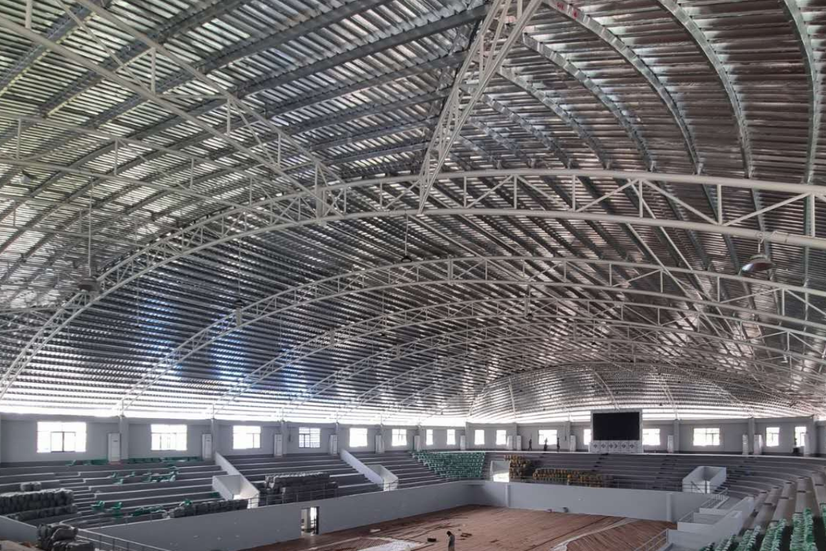 Interior view of the Ba Htoo Indoor Sport Stadium in Mandalay, Myanmar, with the completed steel roof structure and ceiling panels visible above the seating areas.