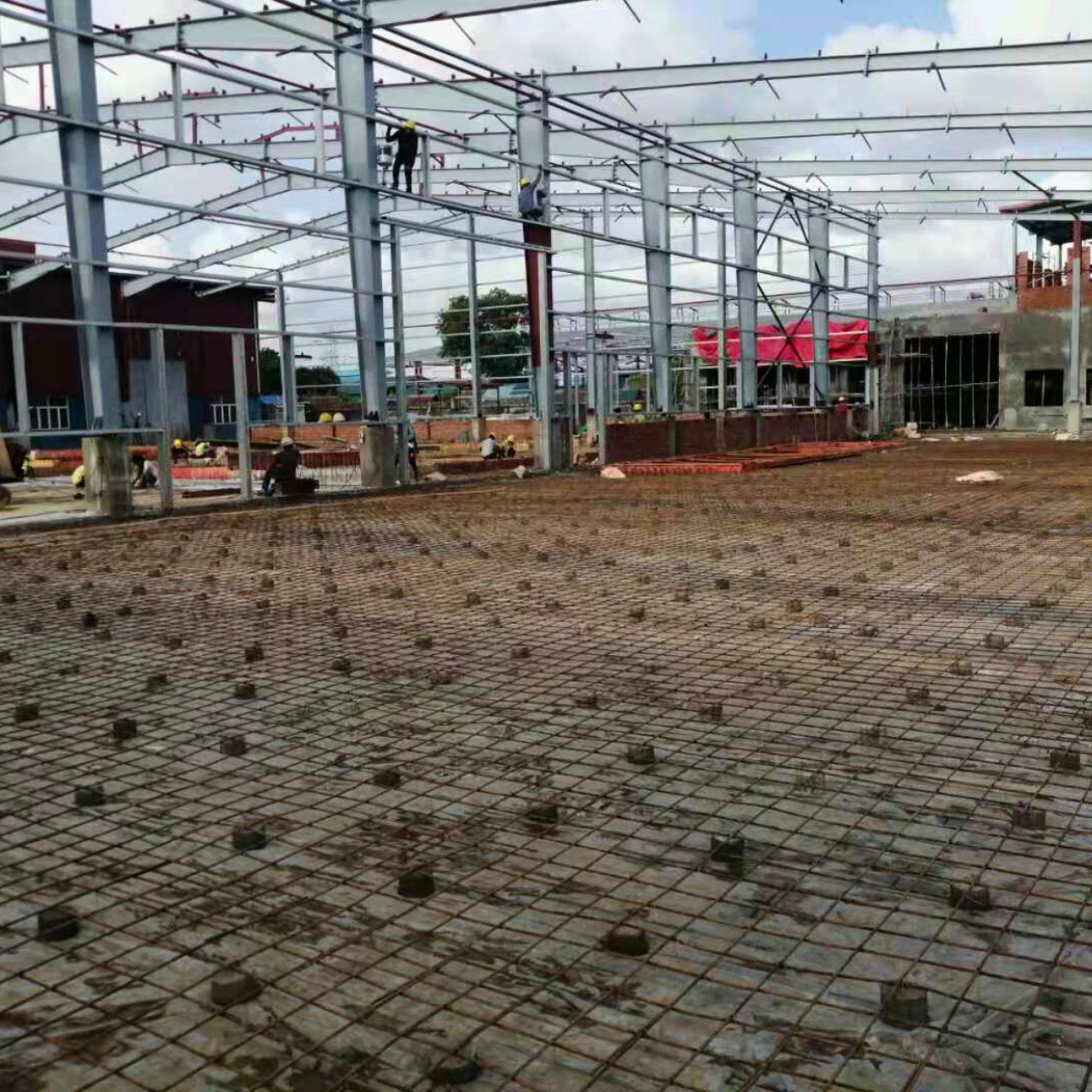 Steel structure warehouse under construction with rebar grid for ground slab and workers installing roof trusses and columns.