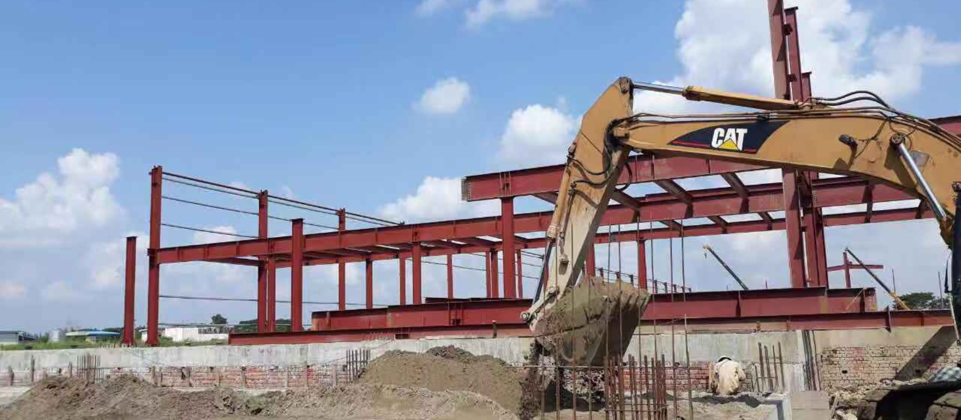 Excavator digging foundation at a steel structure warehouse construction site with red steel frames under a clear sky.