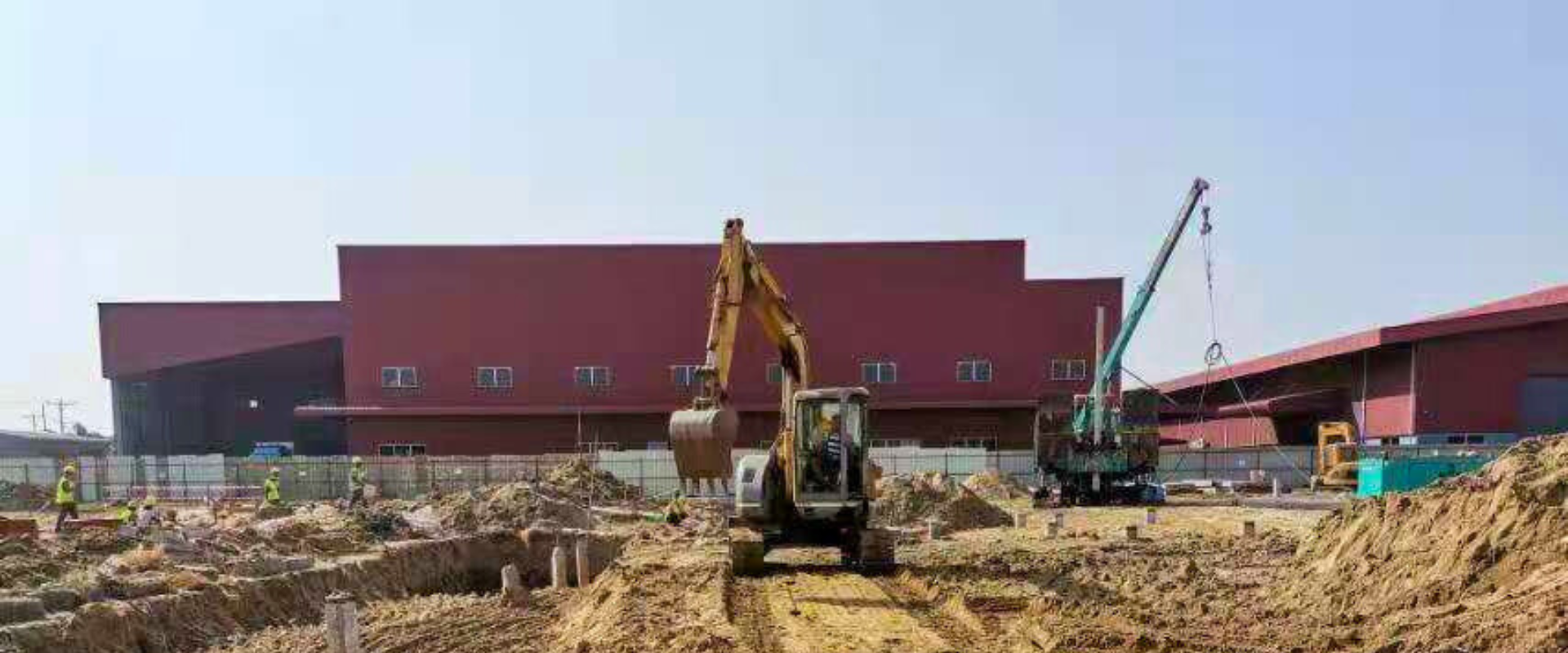 Excavator and crane working on foundation construction for a steel structure warehouse, with workers and equipment on-site in front of a red industrial building.