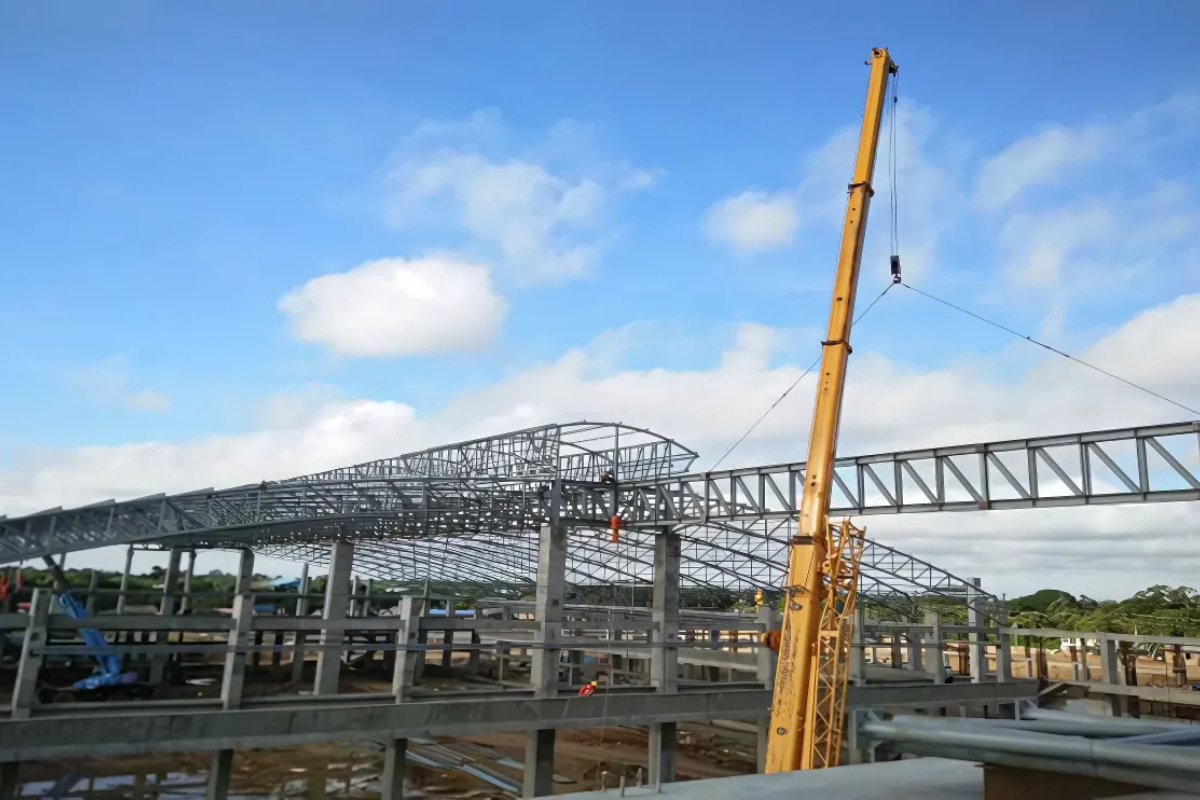 A large yellow crane on the construction site of the Europe & Asia Commercial (EAC) warehouse in Mandalay, actively installing the arc-shaped steel pipe truss roof.