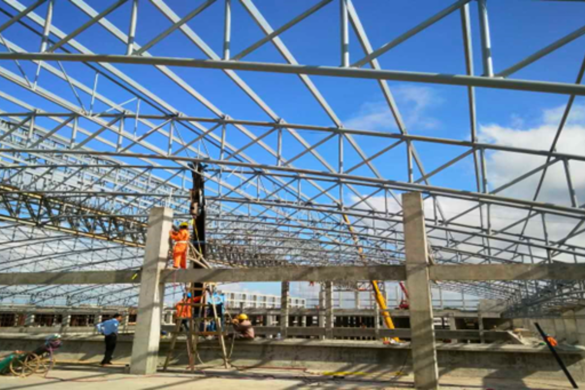 A view of the steel structure construction at the Europe & Asia Commercial (EAC) warehouse in Mandalay, with workers in safety harnesses on the roof truss against a blue sky.