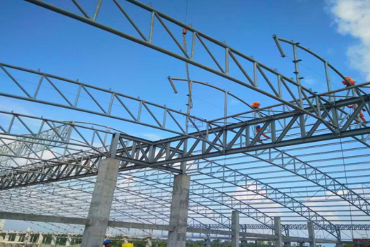 A close-up shot of workers on the steel pipe truss roof of the EAC warehouse project in Mandalay, performing field welding and construction work.