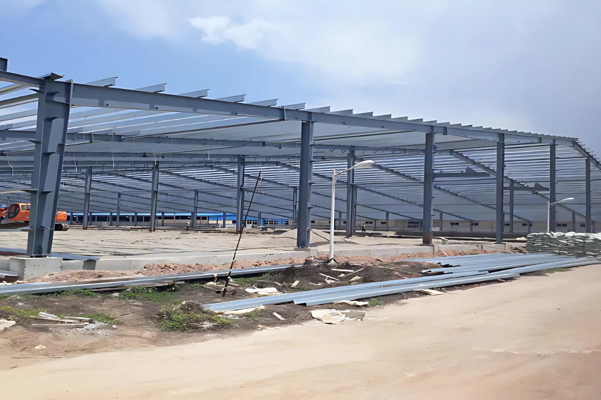 An exterior photo of the Myanmar Knitting Factory in Pathein, showing the steel structure frame being erected and building materials in the foreground.