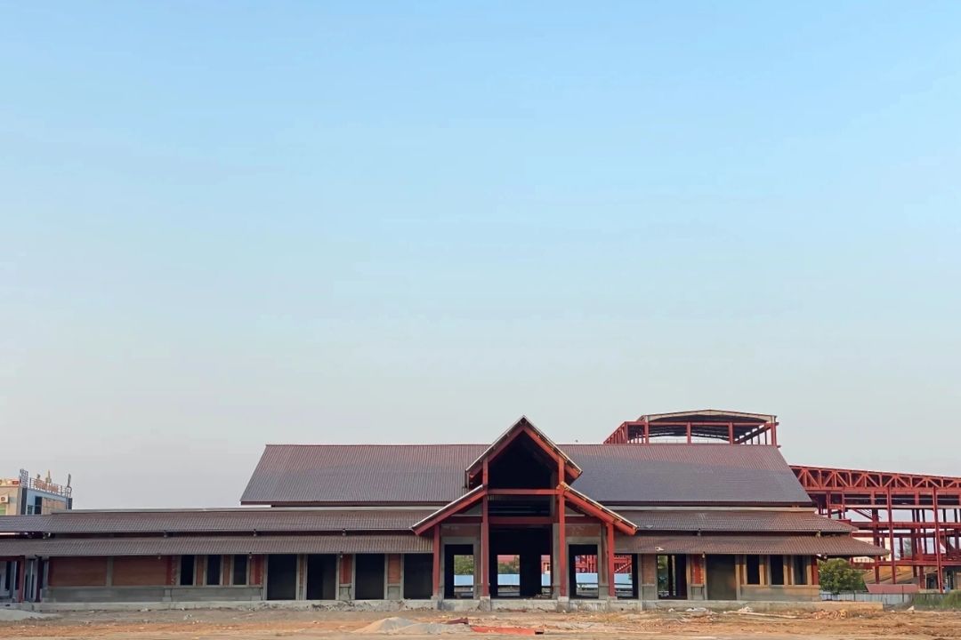 An aerial-like view of the Mandalay Jade Market project by Myanmar Yi Jin, showcasing several buildings in a mid-construction phase with finished roofs and a modern brick-and-concrete facade, set against a backdrop of open fields.