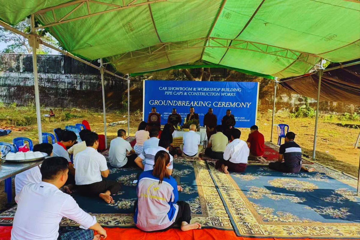 A traditional Buddhist blessing ceremony for the groundbreaking of Performance Auto International’s car showroom and workshop in Myanmar. Monks are seated before an audience of employees on rugs under a green canopy.