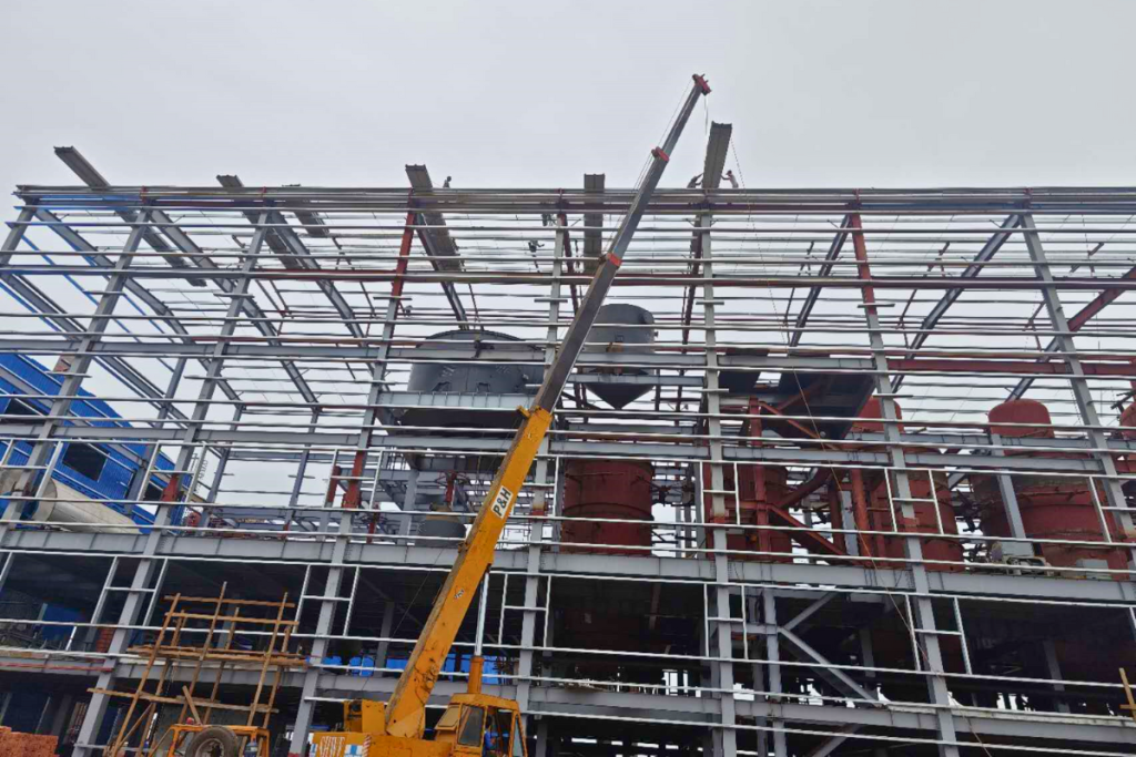 An ongoing industrial construction site by Myanmar Yi Jin featuring an orange mobile crane erecting a multi-storey heavy structural steel framework.
