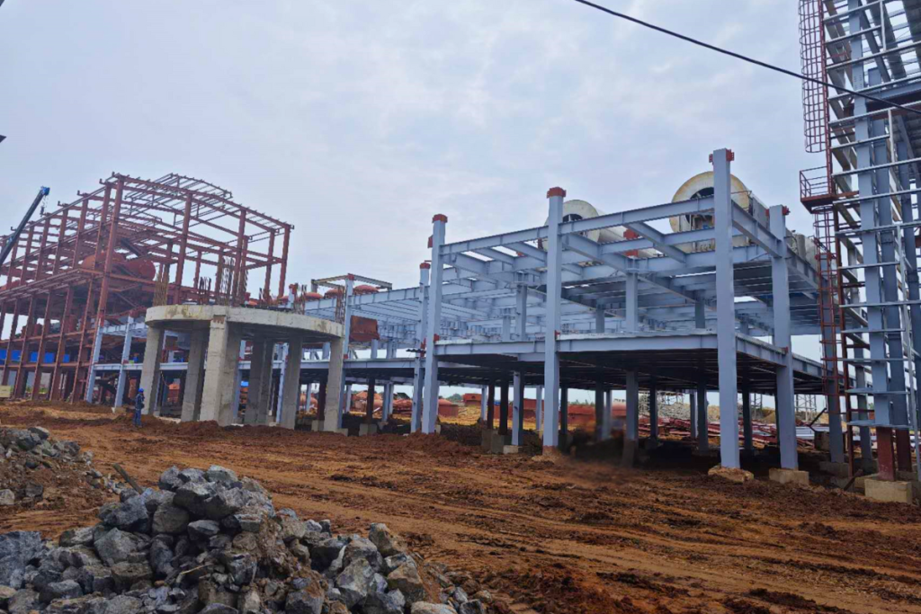 A multi-tiered industrial steel structure building under construction on a red dirt foundation site, showcasing columns and beams installed by Myanmar Yi Jin.