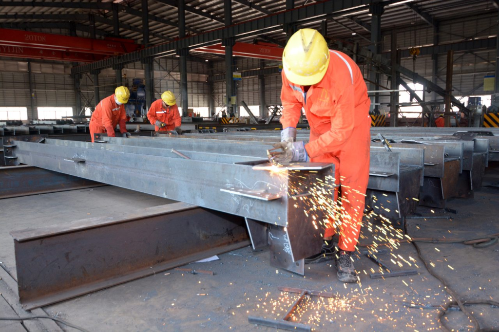 Myanmar Yi Jin factory worker using an angle grinder on a steel structure beam inside the fabrication workshop with sparks flying.